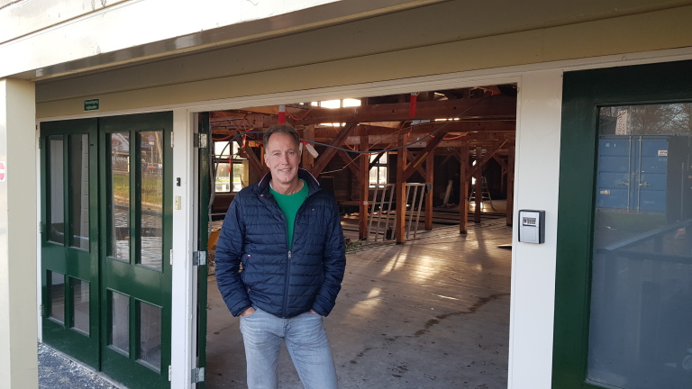 Man in blauwe jas staat bij de ingang van een gebouw met houten balken en een glazen deur.