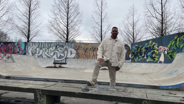 Man in een skatepark met graffiti op de achtergrond, staand met een voet op een betonnen bank.