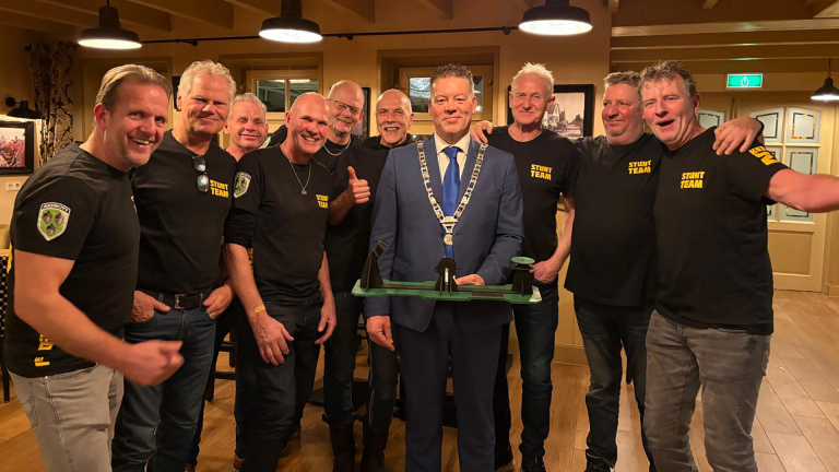 Een groep mannen poseert samen; één man draagt een blauw pak en een ambtsketen, de anderen dragen zwarte T-shirts met "Stunt Team" erop.