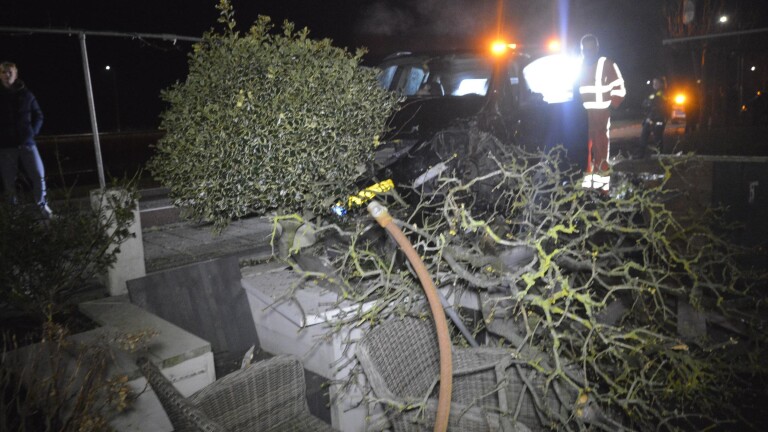 Een auto-ongeluk in het donker waarbij een voertuig tegen een boom en een terras is gebotst; een persoon in veiligheidshesje staat erbij.