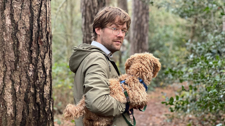 Iemand met een bril draagt een krullende hond in een bos.