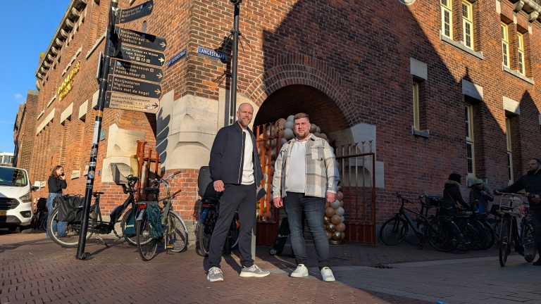 Twee mannen poseren voor een bakstenen gebouw met een boog en straatnaamborden, fietsen en enkele voorbijgangers op de achtergrond.