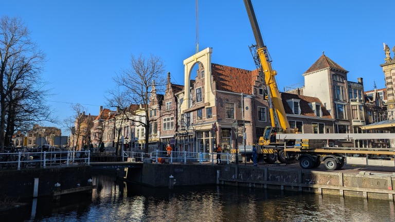 Kraan hijst een grote structuur langs een kanaal met rij van historische gebouwen op de achtergrond onder een heldere blauwe hemel.