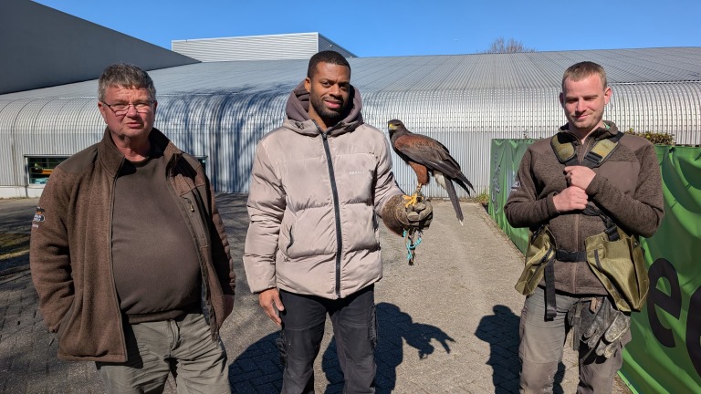 Drie mannen staan buiten naast elkaar; de middelste houdt een roofvogel op zijn hand. Ze hebben allemaal jassen aan. Op de achtergrond is een groot gebouw met een gebogen dak te zien en een groene afzetting. Het is zonnig weer, want er zijn duidelijke schaduwen op de grond.