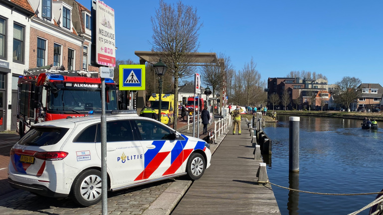 Een politieauto en brandweerwagen staan geparkeerd bij een kade naast een gracht. Er staan mensen, waaronder een brandweerman, op de stoep langs de kade. Een reddingsboot is in het water zichtbaar. Het gebied lijkt onderdeel van een noodgeval of oefening in een stedelijke omgeving.