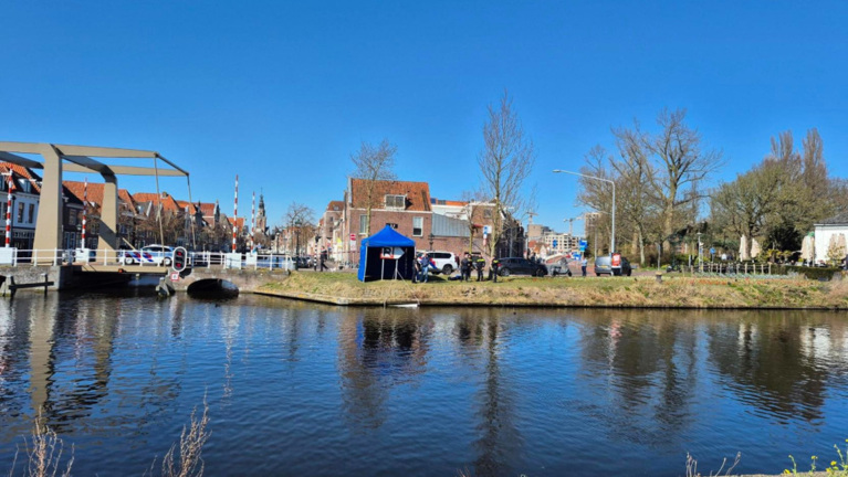 Blauwe tent en politievoertuigen aan de waterkant bij een brug en typisch Nederlandse gebouwen, met een heldere blauwe hemel op de achtergrond.