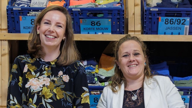 Twee vrouwen glimlachen naar de camera terwijl ze voor planken met opgevouwen kleding in blauwe bakken staan.