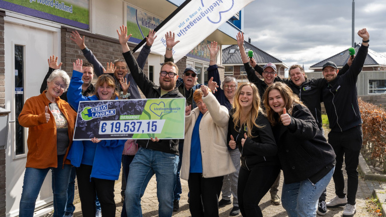 Groep mensen viert buiten met een grote cheque van € 19.537,15. Ze lachen en steken hun handen enthousiast in de lucht. Op de achtergrond zijn een gebouw met een spandoek en enkele huizen te zien.