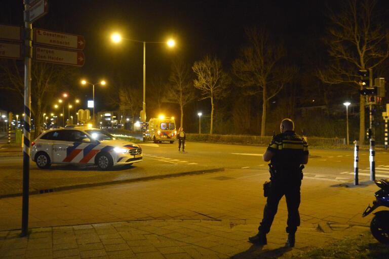 Een Nederlandse politieauto en een ambulance staan geparkeerd op een verlichte weg 's nachts, met een politieagent op de voorgrond en een andere naast de ambulance, en verkeersborden waarop namen van locaties staan aangegeven.