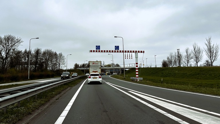 Autos rijden op een weg met een viaduct in de verte, een hoogtebegrenzer staat boven de rijbaan, en er zijn straatlantaarns en bomen langs de berm.