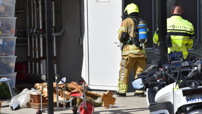 Een brandweerman in beschermende uitrusting staat voor een witte deur, met een politieagent naast hem. Voor hen ligt rommel op de grond, waaronder houten planken en een driewieler. Er staat een politiefiets met apparatuur op de voorgrond.