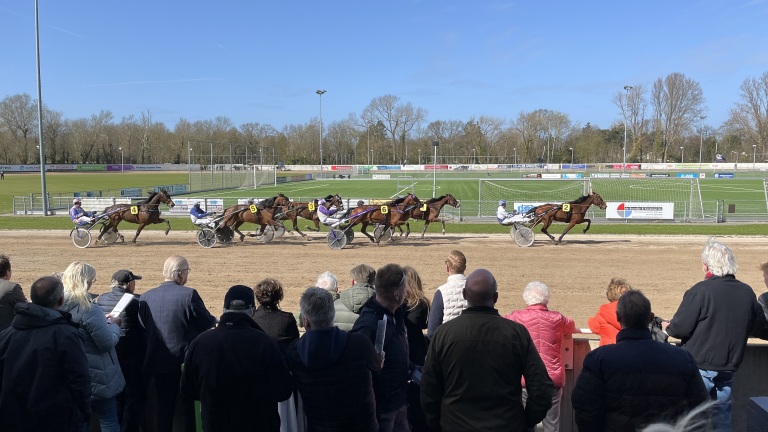 Paardenrennen waarbij zes sulkies racen op een zandparcours, omgeven door toeschouwers die de race bekijken.