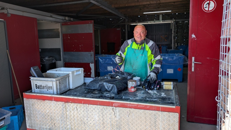 Man in een werkplaats draagt een felgekleurde jas en schort, staat achter een tafel met een doos 'Batterijen', vuilniszakken en andere objecten, en steekt zijn duim omhoog.