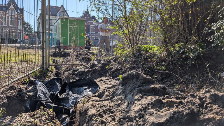 Een bouwplaats met een geul bedekt met zwart plastic, een man op de achtergrond bij een groene container, en omringd door woonhuizen en begroeiing.