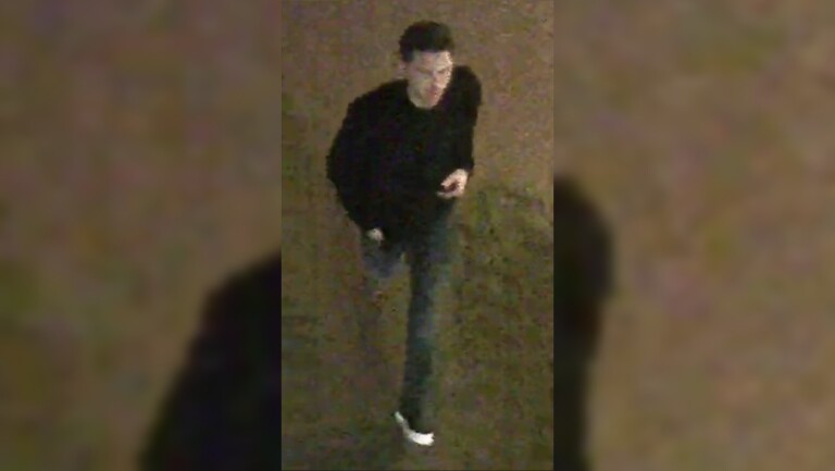 Een persoon loopt over straat in het donker. Ze dragen donkere kleding en witte schoenen.