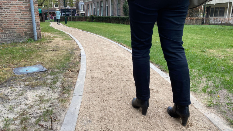 Persoon met zwarte broek en zwarte laarzen loopt over een zandpad naast een grasveld.