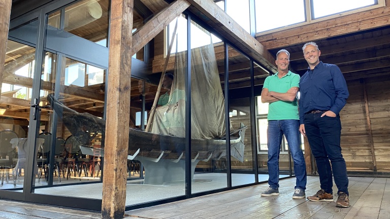 Twee mannen staan naast elkaar in een houten gebouw met een glazen wand, waarachter een traditionele houten boot met een opgetuigd zeil te zien is.