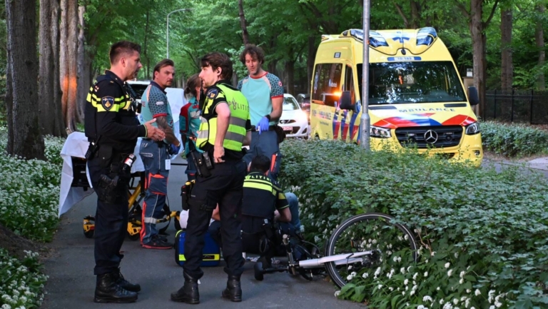 Politie en medisch personeel verlenen hulp bij een fietsongeval in een bosrijke omgeving, met een ambulance op de achtergrond.