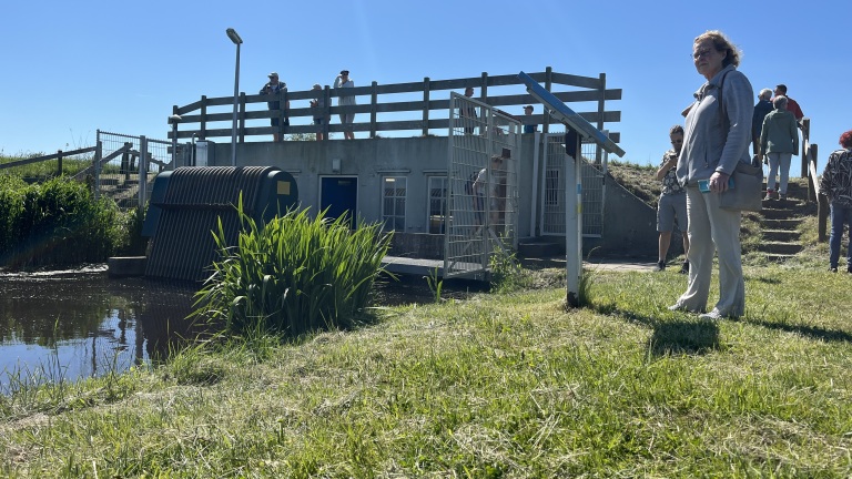 Groep mensen staat buiten bij een gebouw met een hek en trap, omringd door gras en water, onder een heldere blauwe hemel.