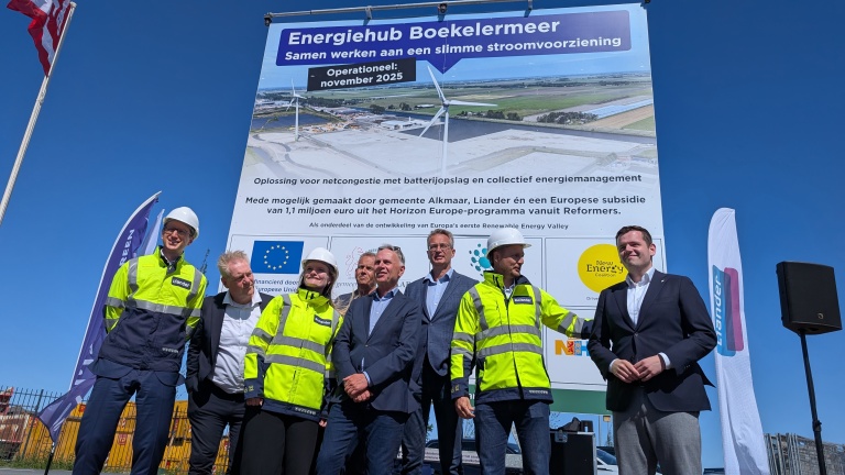 Groep mensen poseert voor een groot bord met informatie over Energiehub Boekelermeer, met tekst en afbeeldingen van windturbines, op een zonnige dag.