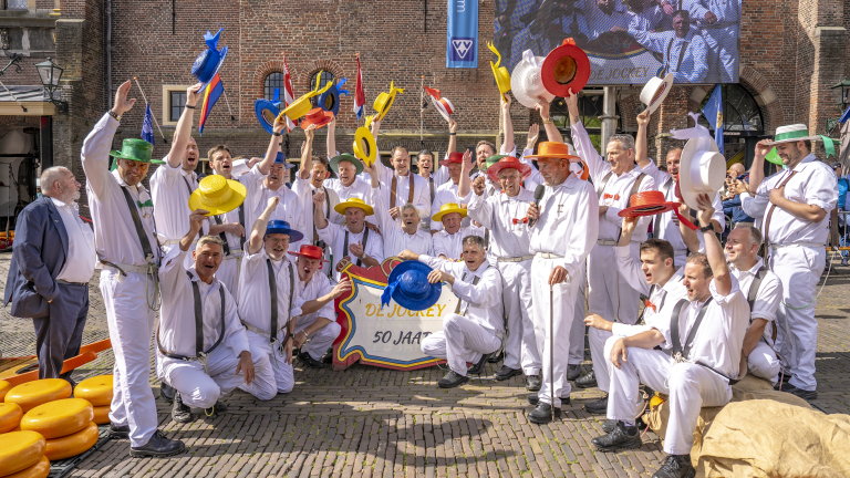 Een groep mensen in witte kleding viert feest op een plein met kleurrijke hoeden in hun handen. Op de achtergrond is een historisch gebouw en een bord met '50 Jaar' zichtbaar.