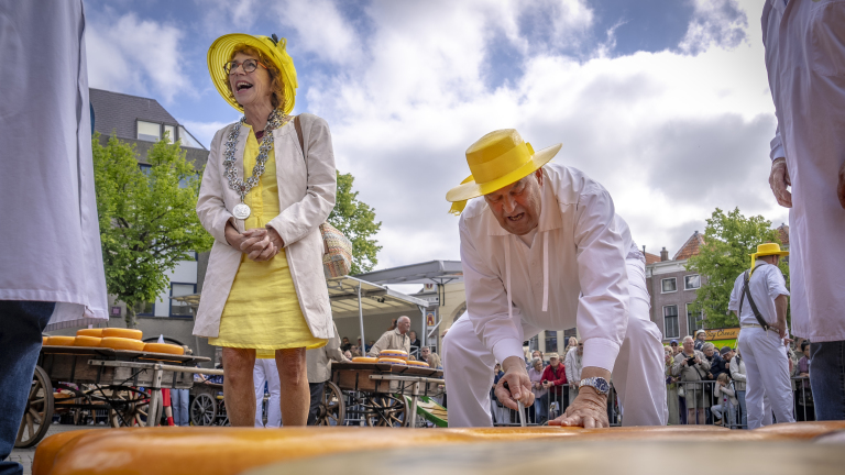 Twee personen in gele hoeden bij een kaasmakt, één van hen hakt in een grote kaas.