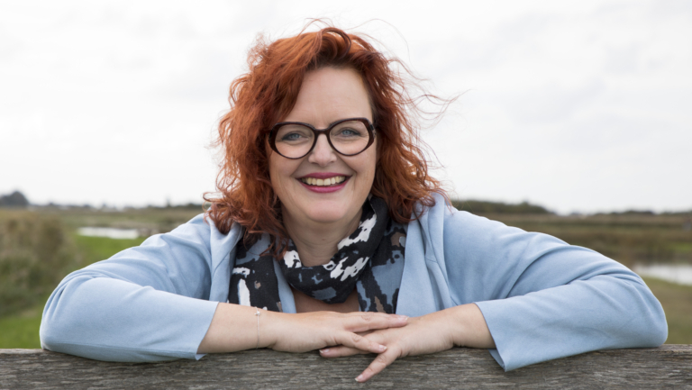 Vrouw met rood haar en een bril, leunend aan de rand van een houten hek, in een open veld met een rivier op de achtergrond.