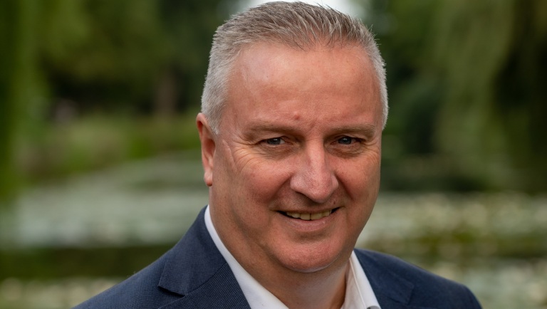Portret van een glimlachende man in een pak, met een groene, onscherpe achtergrond.