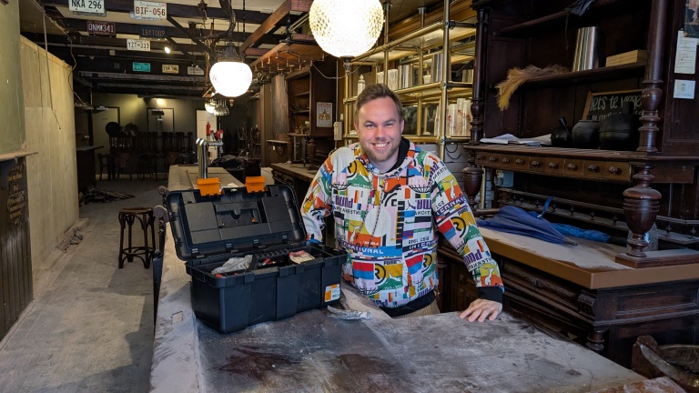 Een man in een kleurrijke trui staat glimlachend achter een werkbank met een gereedschapskoffer in een stoffige werkplaats, met ouderwetse meubels en plafondverlichting zichtbaar op de achtergrond.