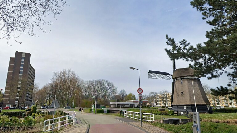 Straatbeeld met een traditionele Hollandse windmolen aan de rechterkant en een hoog flatgebouw aan de linkerkant, omgeven door groene bomen en struiken. Er is een fietspad met een bruggetje en enkele verkeersborden op de voorgrond. De lucht is licht bewolkt.