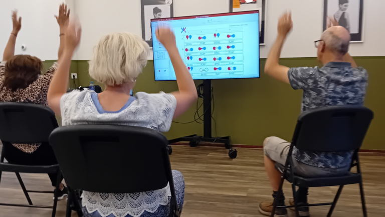 Drie mensen zitten op stoelen en strekken hun armen naar boven terwijl ze naar een scherm kijken dat instructies toont in een klaslokaal.