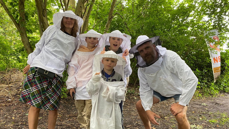 Een groep mensen, waaronder kinderen, dragen bijenkostuums met beschermende hoeden in een bosrijke omgeving.