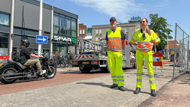 Twee mensen in fluorescerende werkkleding staan voor een vrachtwagen op een straat naast een Spar supermarkt, terwijl een man op een motorfiets voorbijrijdt.