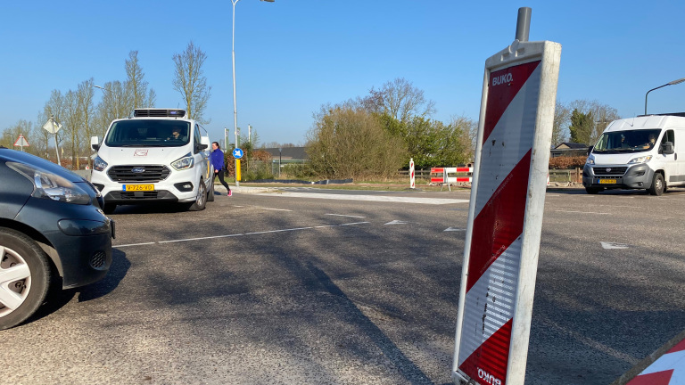 Een kruising met een wegwerkzaamheden bord op de voorgrond, een witte bestelwagen en een vrouw die de straat oversteekt, omgeven door bomen en een heldere blauwe lucht.