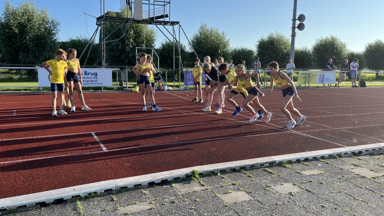 Kinderen in sportkleding staan klaar om te rennen op een atletiekbaan op een zonnige dag, met toeschouwers in de achtergrond.