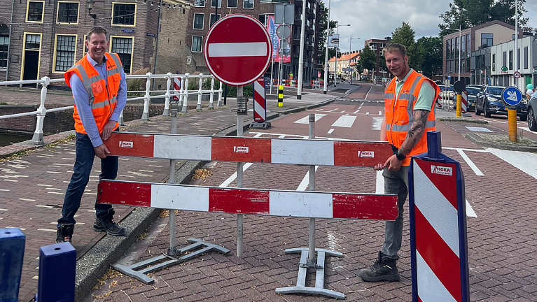 Twee mannen in oranje veiligheidsvesten verplaatsen een rood-wit afzethek op een straat in een stedelijke omgeving.