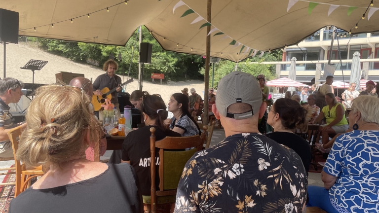 Groep mensen zit onder een tentluifel bij een muziekoptreden met een gitarist op de achtergrond.