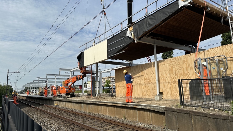 Constructiewerkzaamheden aan een station, waar arbeiders in oranje kleding bezig zijn met het plaatsen van een metalen overkapping met behulp van hijskranen boven een perron.