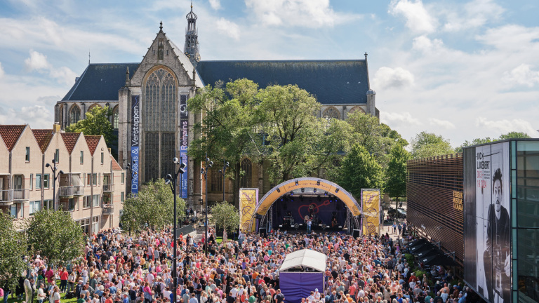 Een drukbezocht openluchtconcert voor de Grote Kerk van Breda, met een podium en een groot aantal toeschouwers op een zonnige dag.