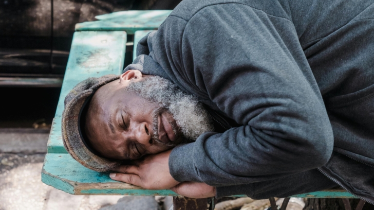 Man sleeping on a bench in a city, next to a cardboard sign reading "WILL WORK FOR FOOD" and a paper cup on the ground.