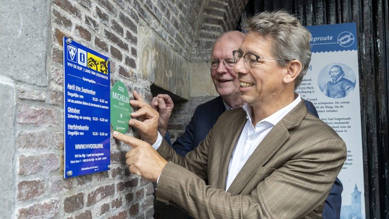 Twee mannen in pakken bevestigen een groene Michelin-gids sticker voor 2025 op een bakstenen muur naast een blauw bord met openingsuren in tekst.