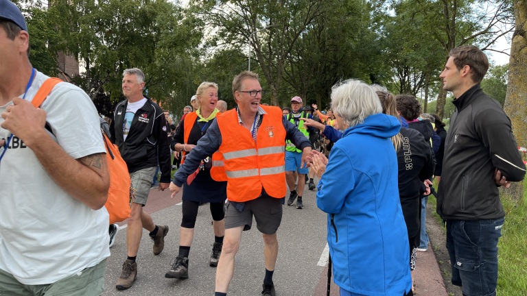 Mensen lopen in een groep over een weg, met één persoon in een fel oranje veiligheidsvest die lachend contact maakt met toeschouwers langs de kant. Er zijn meerdere mensen in sportieve kleding en verschillende achter hen marcherende figuren, tegen een achtergrond van bomen en een heldere lucht.