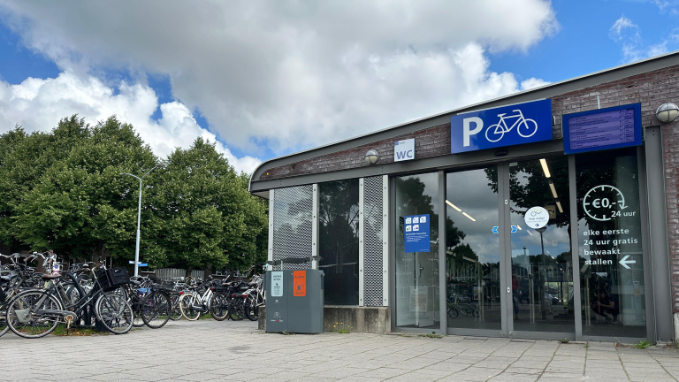 Fietsenstalling met meerdere fietsen geparkeerd naast een gebouw met glazen deuren, waarop een bord met een witte fiets op een blauwe achtergrond hangt en prijsaanduidingen te zien zijn. Er staan ook afvalbakken naast het gebouw en op de achtergrond zijn groene bomen en bewolkte lucht zichtbaar.