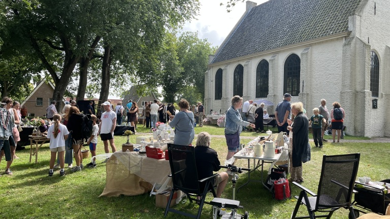 Een buitenmarkt met mensen die rondlopen en staan bij tafels met tweedehands goederen, naast een witte kerk met hoge ramen.