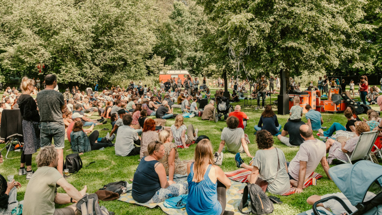 Een grote groep mensen zit en staat op het gras in een park, luisterend naar een live muziekoptreden onder de bomen.