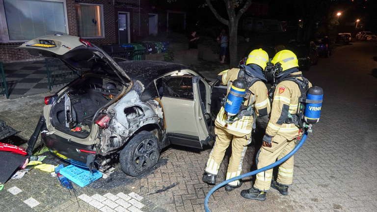 Twee brandweerlieden in uniform inspecteren een uitgebrande auto met open kofferruimte, staande op een straat bij nacht.