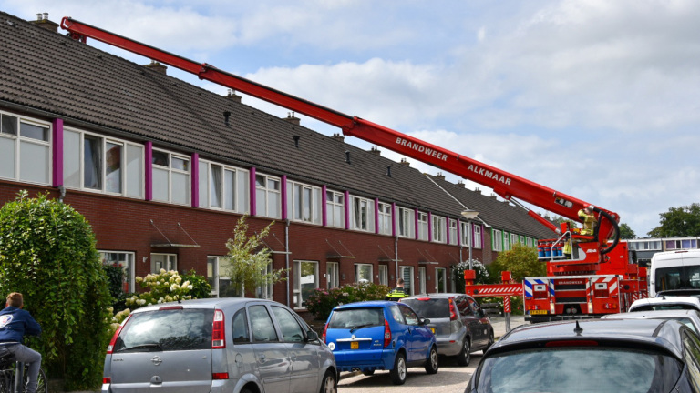 Een brandweerwagen met een lange rode ladder van de brandweer Alkmaar, geparkeerd voor een rijtjeshuis. Een brandweerman staat op het platform van de ladder die naar het dak van het huis reikt. Er staan auto's geparkeerd langs de straat en een kind op een fiets kijkt naar de actie.