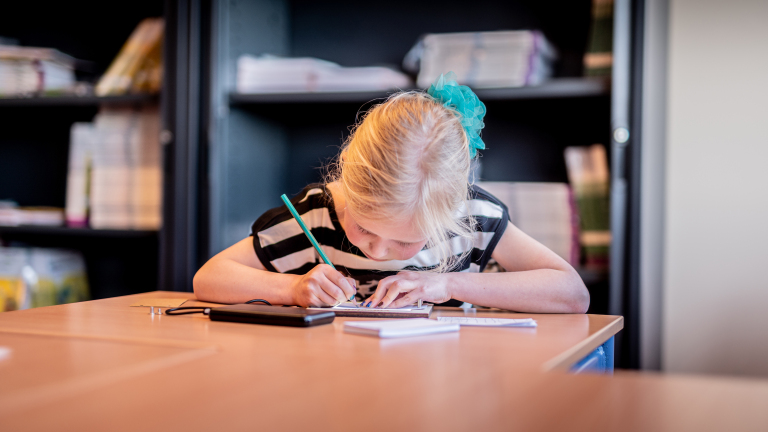 Een jong meisje met een blauw lint in haar haar schrijft geconcentreerd met een potlood in een notitieblok op een tafel, in een ruimte met boekenplanken op de achtergrond.