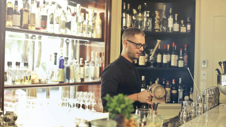 Een barman in een zwart hemd en bril die dranken inschenkt achter een bar, omgeven door een grote selectie flessen sterke drank op planken.