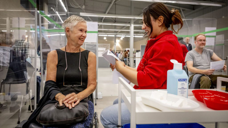 Een lachende oudere vrouw zit met een jas over haar arm tegenover een jonge vrouw in een rood uniform die een formulier vasthoudt in een vaccinatielocatie. Op de voorgrond is een tafel te zien met ontsmettingsmiddel en medische benodigdheden.